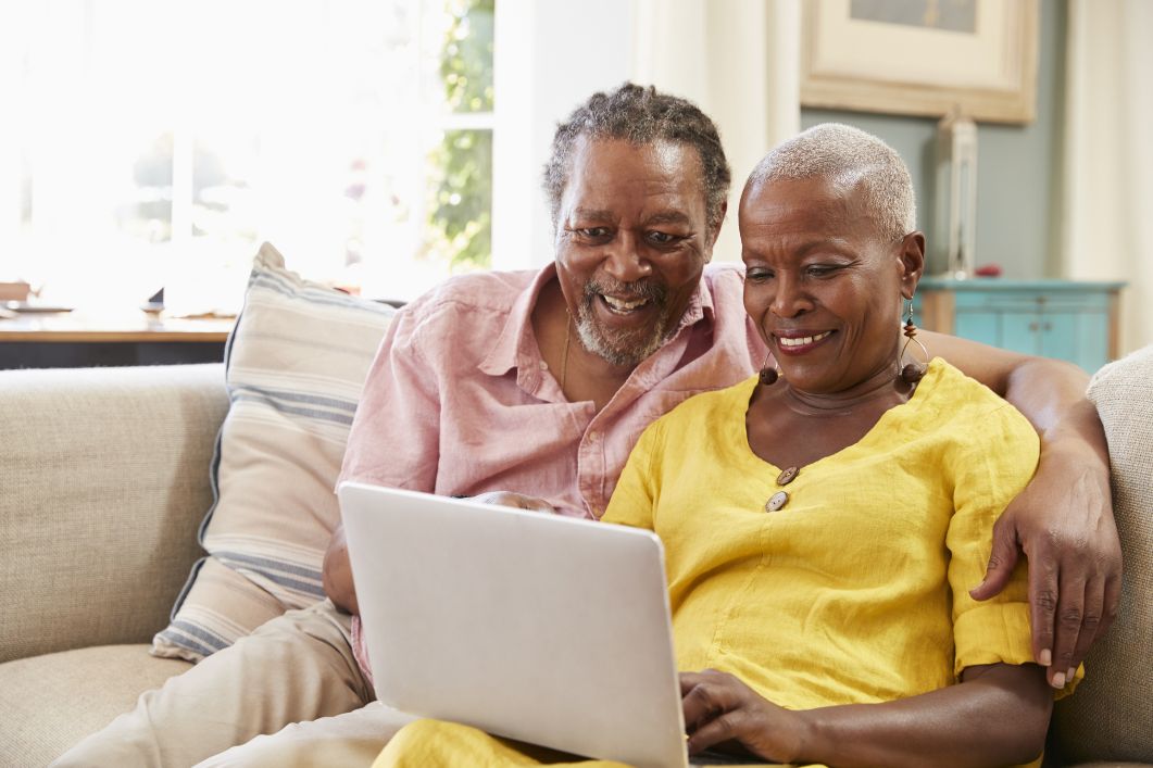 an elderly man has his arm lovingly over an elderly woman's shoulder while they both smile at a computer screen while selling an inherited house